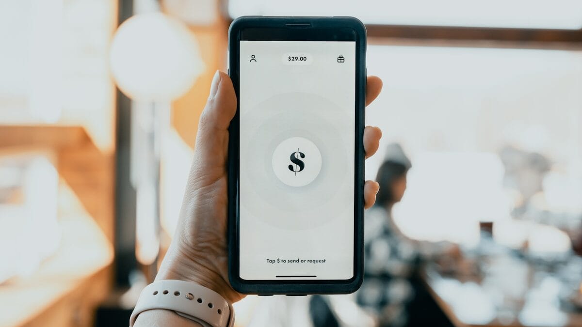 A hand holds a smartphone displaying a money transfer app with a 29.00 balance in a blurred indoor setting.