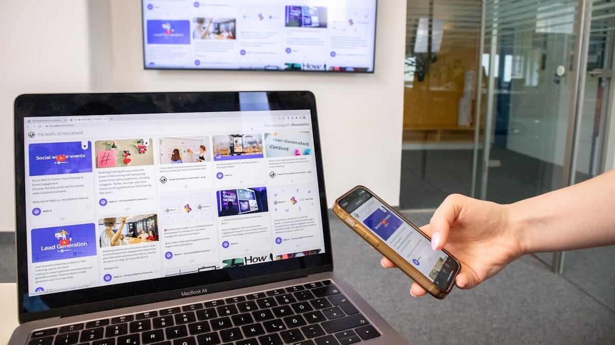 A person holds a smartphone while viewing a laptop and a wall-mounted screen, both displaying similar website content.