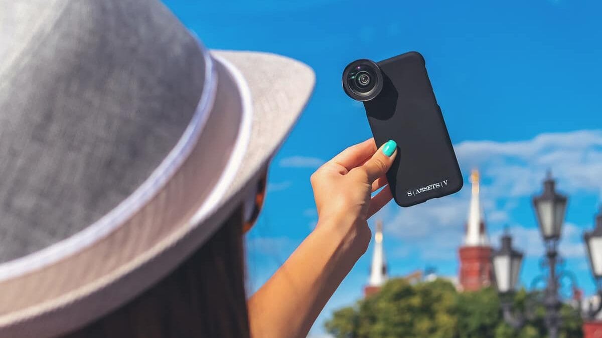 Person wearing a hat holds up a smartphone with an external lens attached, with a cityscape and blue sky in the background.