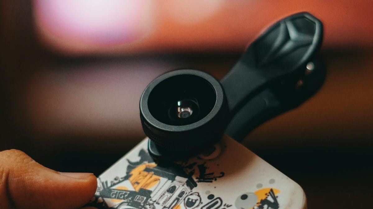 A hand holds a smartphone with a black clip-on lens attached over a case featuring black-and-white graffiti-style artwork.