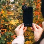 Person holding a smartphone with an external lens attachment, taking a photo of colorful autumn foliage.