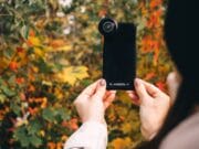 Person holding a smartphone with an external lens attachment, taking a photo of colorful autumn foliage.