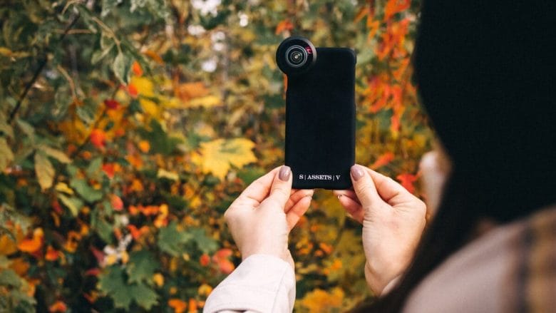 Person holding a smartphone with an external lens attachment, taking a photo of colorful autumn foliage.