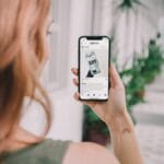 A person with reddish hair holds a smartphone, browsing a social media post, while standing in a corridor with potted plants.