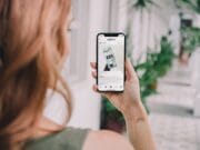 A person with reddish hair holds a smartphone, browsing a social media post, while standing in a corridor with potted plants.