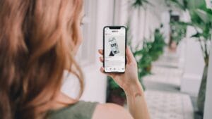 A person with reddish hair holds a smartphone, browsing a social media post, while standing in a corridor with potted plants.