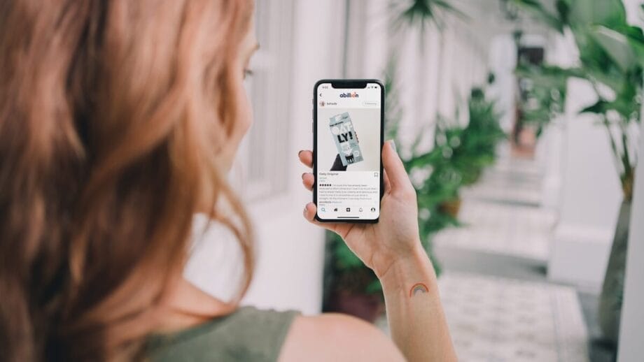 A person with reddish hair holds a smartphone, browsing a social media post, while standing in a corridor with potted plants.