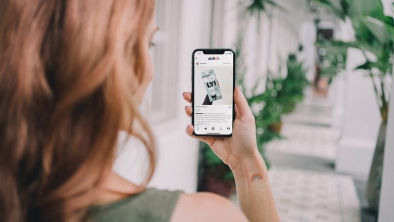 A person with reddish hair holds a smartphone, browsing a social media post, while standing in a corridor with potted plants.