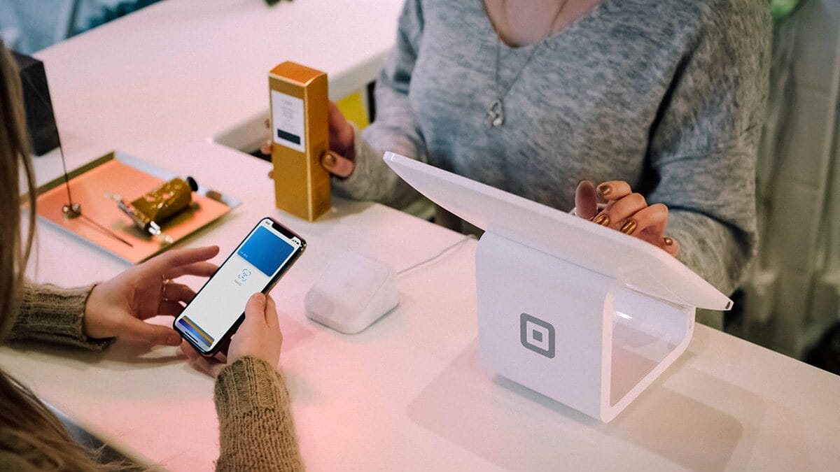 A person holds a phone to make a contactless payment at a white Square register while another person stands behind the counter.