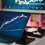 A tablet displays a candlestick stock chart on a desk with a keyboard, calculator, pen, and computer monitors in the background.