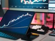 A tablet displays a candlestick stock chart on a desk with a keyboard, calculator, pen, and computer monitors in the background.