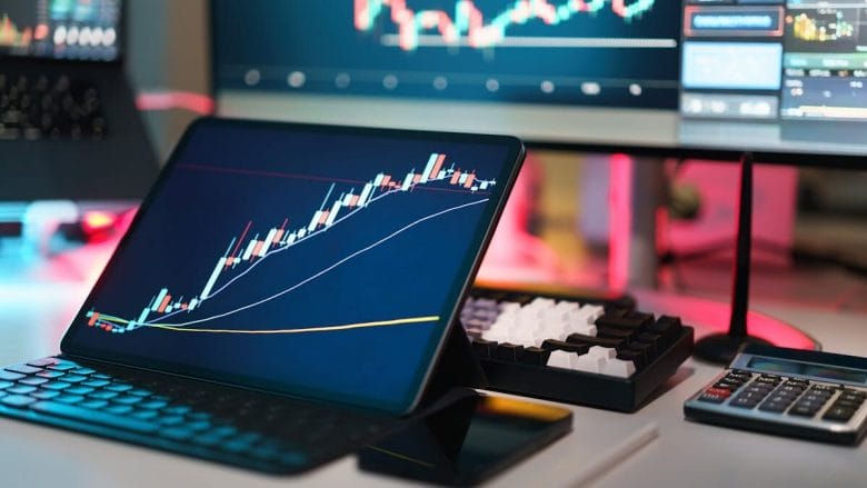 A tablet displays a candlestick stock chart on a desk with a keyboard, calculator, pen, and computer monitors in the background.