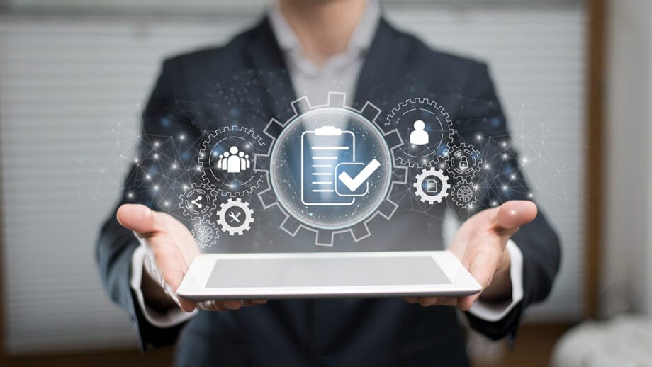 A person in a suit holds a tablet with digital icons of gears, a checklist, and people superimposed above it, representing task management.