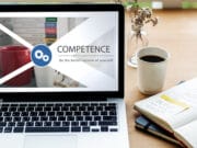 A laptop displays the word COMPETENCE and Be the better version of yourself beside a mug, books, and an open notebook on a desk.