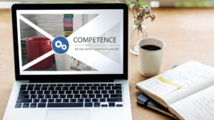 A laptop displays the word COMPETENCE and Be the better version of yourself beside a mug, books, and an open notebook on a desk.