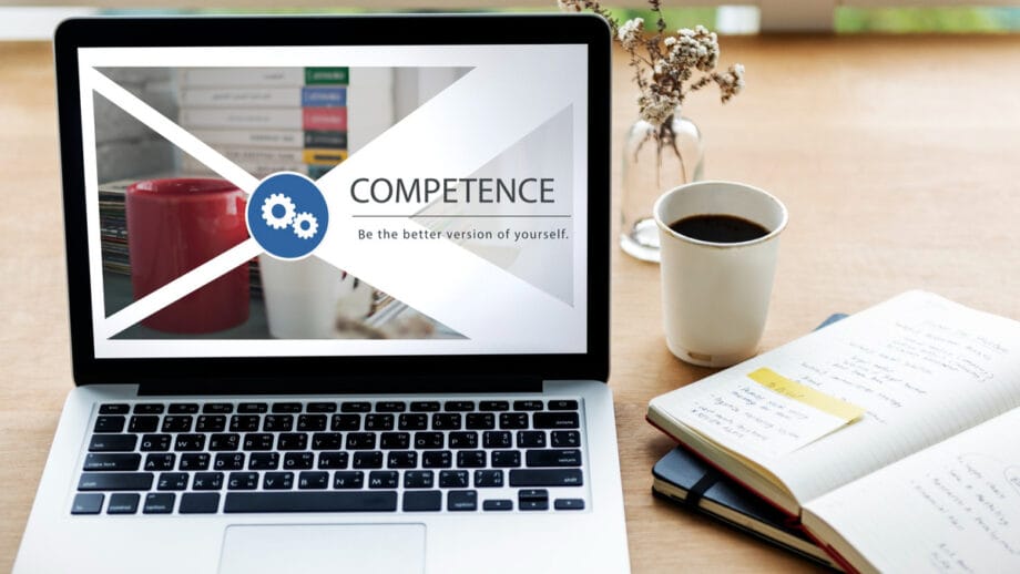 A laptop displays a AI Document Verification template with the word COMPETENCE and Be the better version of yourself beside a mug, books, and an open notebook on a desk.