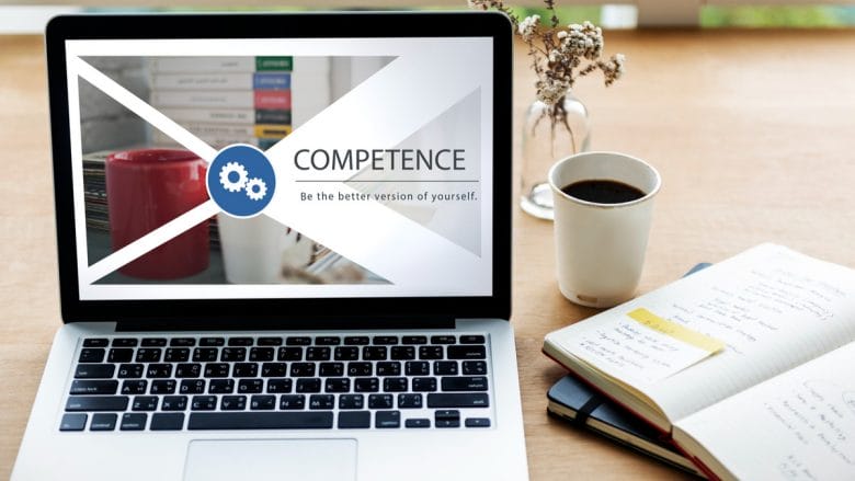 A laptop displays the word COMPETENCE and Be the better version of yourself beside a mug, books, and an open notebook on a desk.