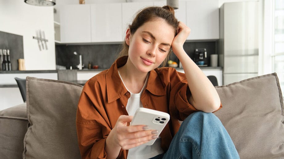 Woman sitting on a couch in a modern kitchen, looking at her smartphone with a relaxed expression.