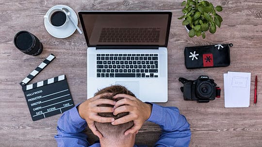 A person sits at a desk with a laptop, camera, coffee, and film clapperboard, holding their head in frustration.