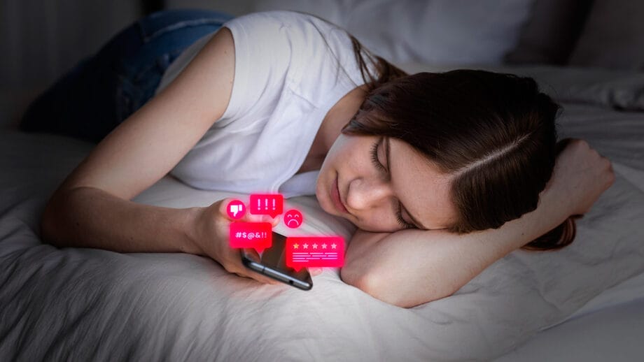 A young woman lies on a bed looking at her phone, which displays negative emoji reactions and angry messages.