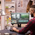 A man in a plaid shirt edits video on a computer at a modern desk with a lamp, plant, and office supplies; a woman stands in the background.