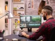 A man in a plaid shirt edits video on a computer at a modern desk with a lamp, plant, and office supplies; a woman stands in the background.
