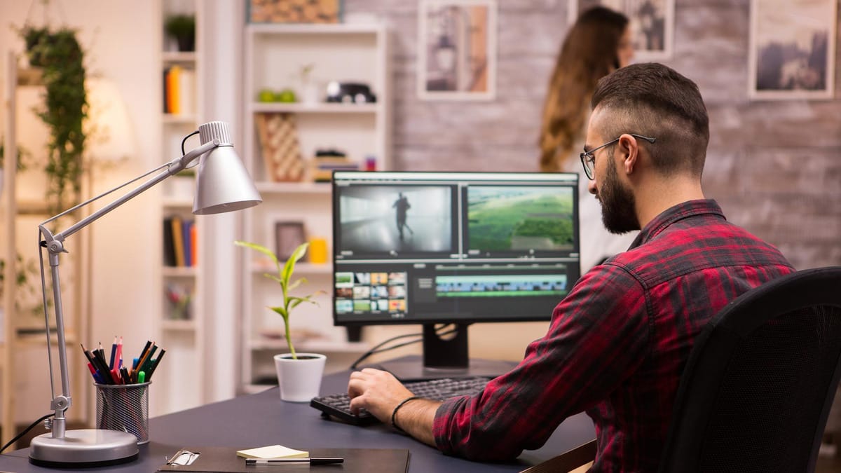 A man in a plaid shirt edits video on a computer at a modern desk with a lamp, plant, and office supplies; a woman stands in the background.