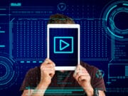 A person holds a tablet in front of their face displaying a play button, with digital graphic elements in the background.