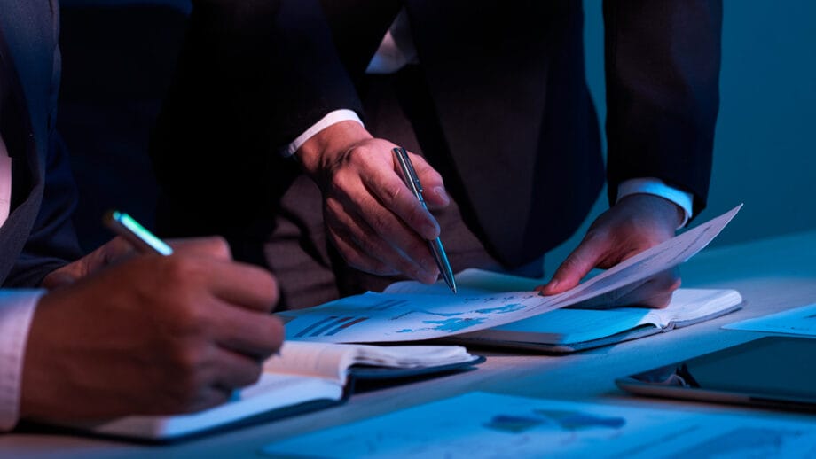 Two people in business attire reviewing documents and taking notes at a desk with charts and a tablet under blue lighting.