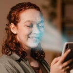 A woman uses facial recognition on her smartphone while holding a mug and smiling.