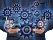 Person in a suit holding a tablet with digital gears and cogs overlayed, representing technology and process automation.