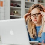 Woman wearing headphones and glasses sits at a desk with a laptop, touching her temples and appearing stressed or frustrated.