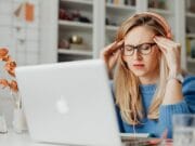 Woman wearing headphones and glasses sits at a desk with a laptop, touching her temples and appearing stressed or frustrated.