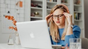 Woman wearing headphones and glasses sits at a desk with a laptop, touching her temples and appearing stressed or frustrated.