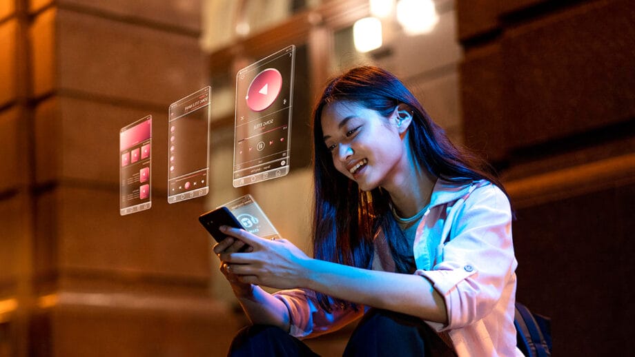 A woman sitting outdoors at night uses her smartphone while digital music app interfaces are projected in front of her.