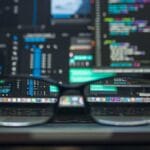 A pair of eyeglasses rests on a laptop keyboard with programming code displayed on the laptop screen in the background.