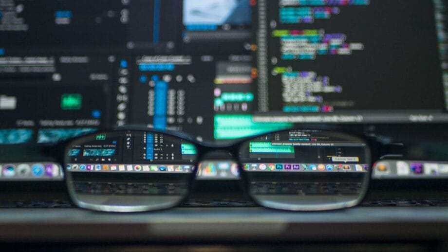 A pair of eyeglasses rests on a laptop keyboard with programming code displayed on the laptop screen in the background.
