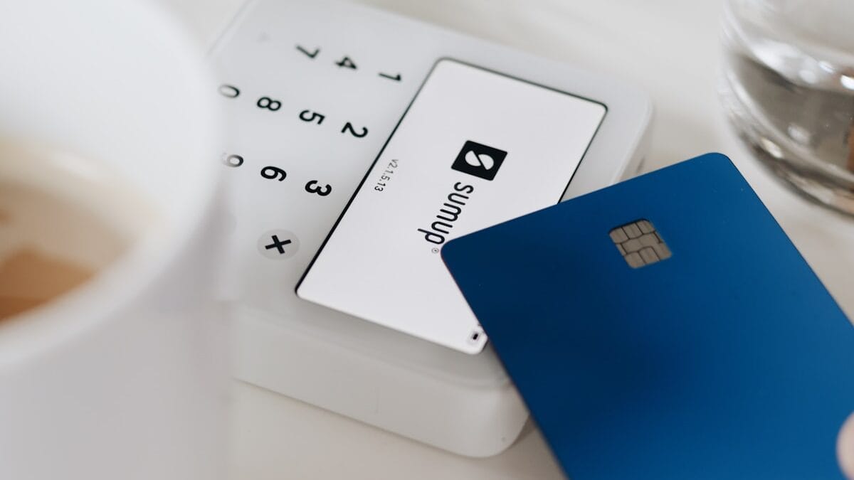 A blue payment card is held near a white SumUp card reader on a table next to a cup and a glass of water.