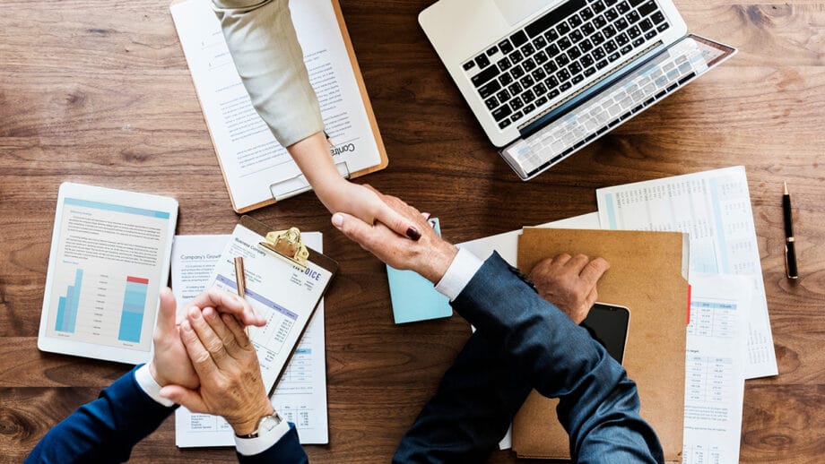 Two people shake hands over a wooden desk with a laptop, charts, documents, and a clipboard, indicating a business meeting or agreement.