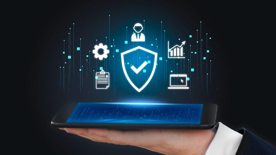 A hand holds a tablet displaying digital security icons, including a shield with a checkmark, gears, graphs, and a clipboard.
