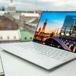A slim white laptop on a table displays a weather app for Paris, France, with city buildings visible through a window in the background.