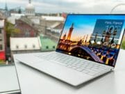 A slim white laptop on a table displays a weather app for Paris, France, with city buildings visible through a window in the background.