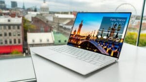 A slim white laptop on a table displays a weather app for Paris, France, with city buildings visible through a window in the background.