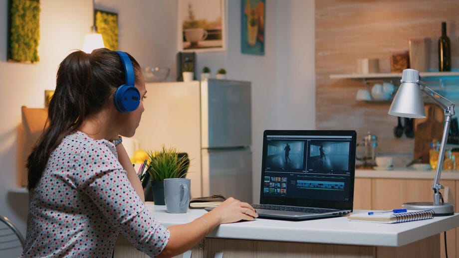 A woman wearing headphones edits video on a laptop at a desk in a kitchen, with a mug, notebook, and lamp beside her.