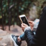 Person sitting on outdoor steps, holding and looking at a smartphone, with greenery and a railing in the background.