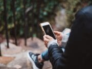 Person sitting on outdoor steps, holding and looking at a smartphone, with greenery and a railing in the background.