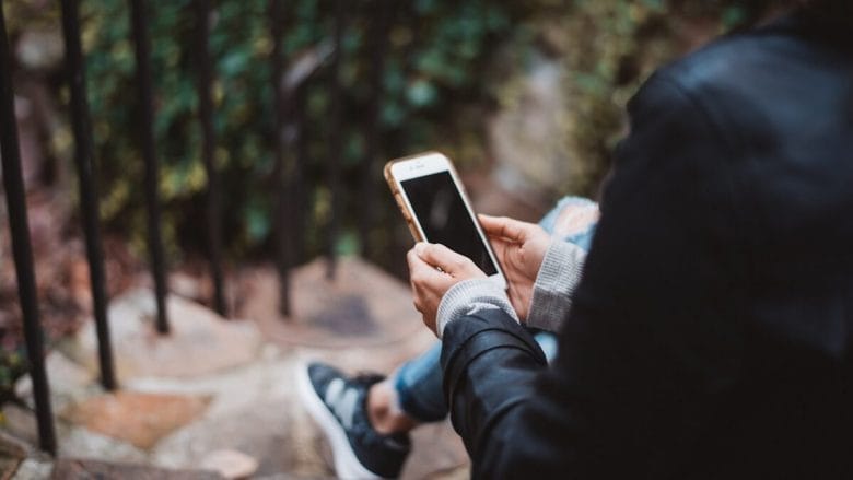 Person sitting on outdoor steps, holding and looking at a smartphone, with greenery and a railing in the background.