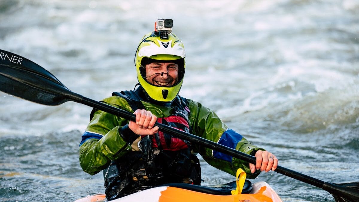 A person in a helmet and waterproof gear kayaks in rough water, holding a paddle and wearing a GoPro camera.