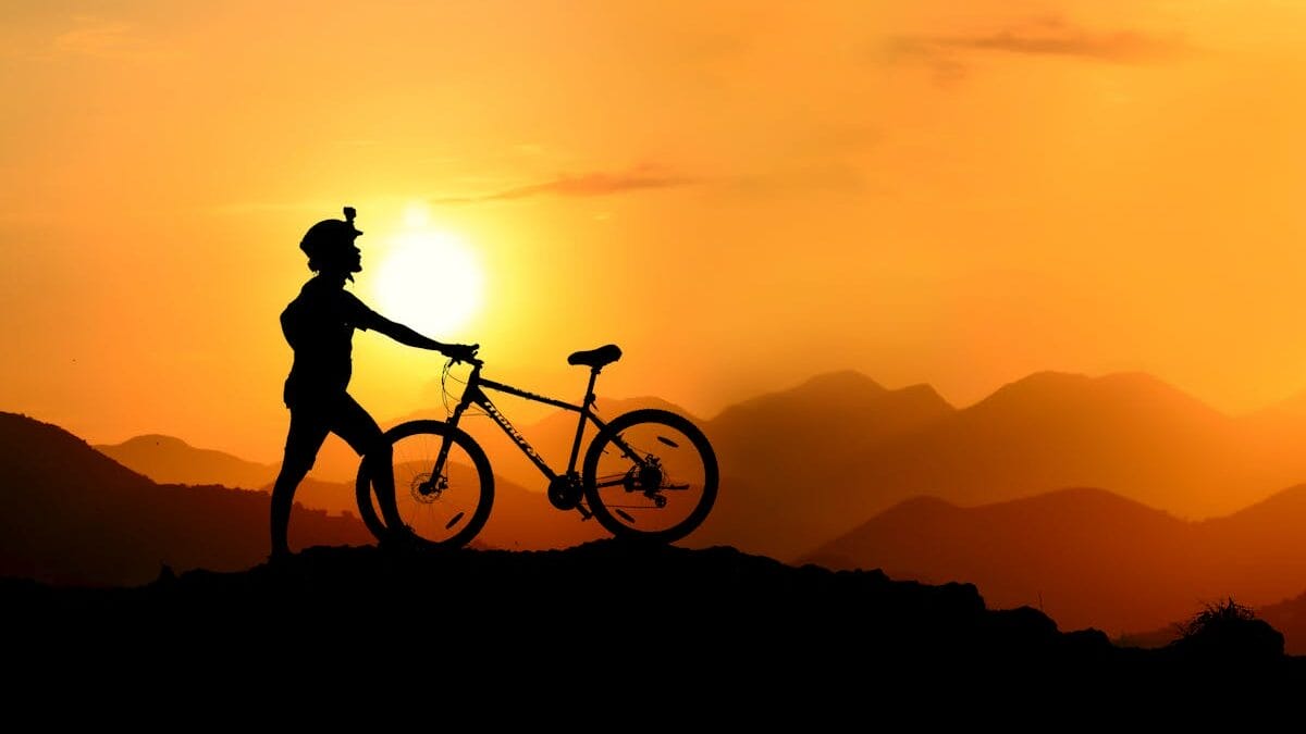 Silhouette of a person standing with a mountain bike on a ridge at sunset, with mountains and a bright sky in the background.