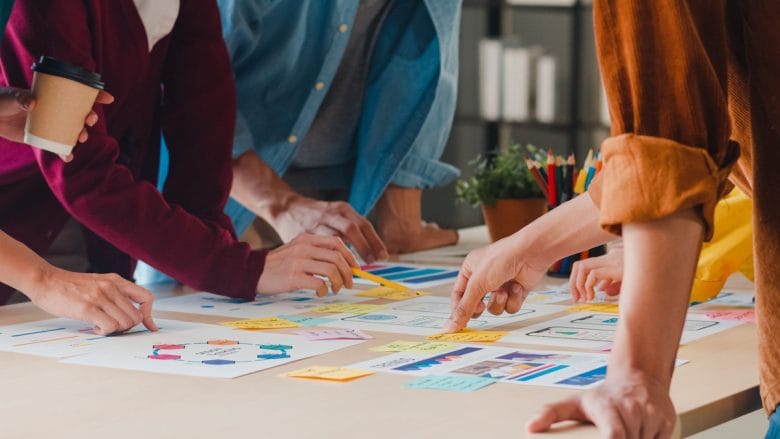 Several people stand around a table, pointing at charts and sticky notes during a collaborative planning or brainstorming session.
